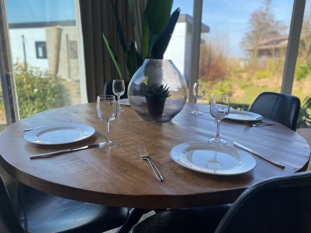 Eetkamer met ronde houten tafel, glazen vaas met plant, servies en bestek voor vier personen, uitzicht op groene omgeving