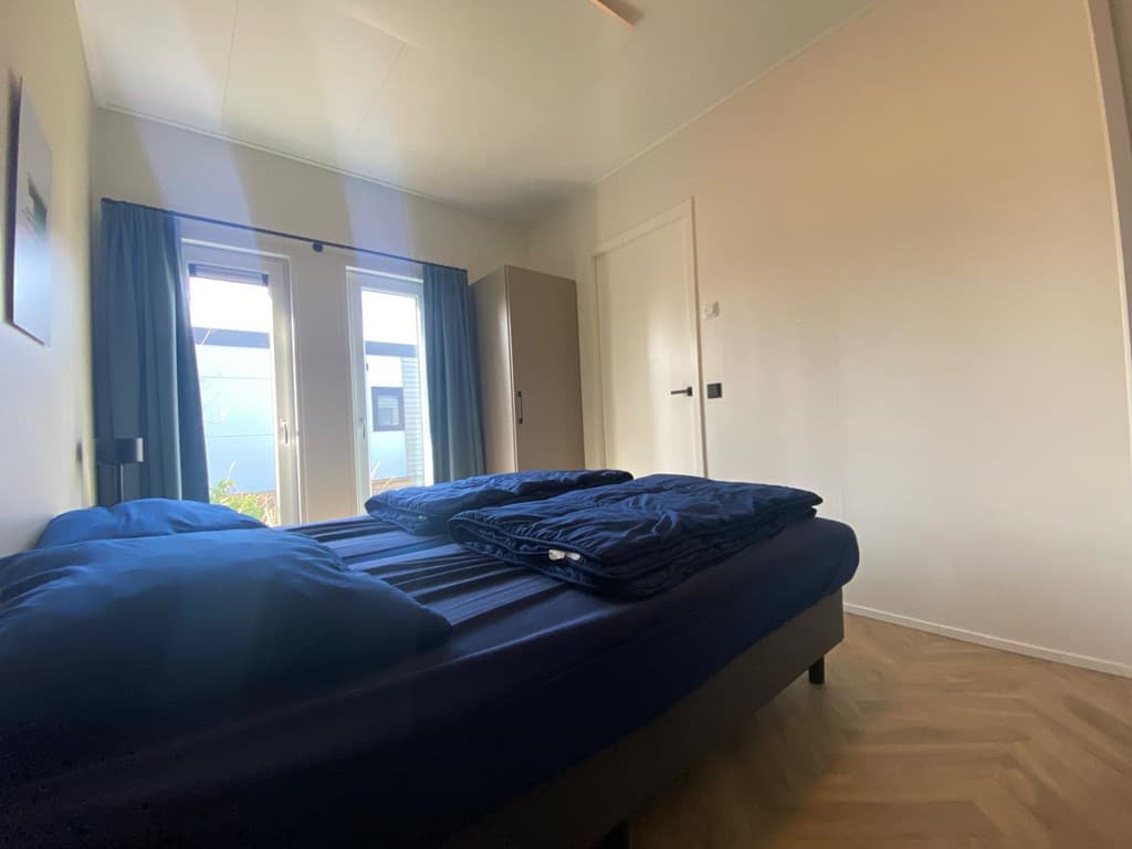 Tweepersoonsbed met blauwe dekbedden en toegang tot buiten via dubbele deuren in een lichte slaapkamer