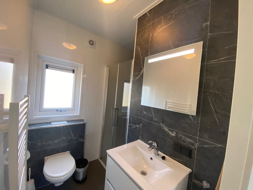 Moderne badkamer met witte en grijze tegels, douchecabine, wastafel met spiegel en toilet.