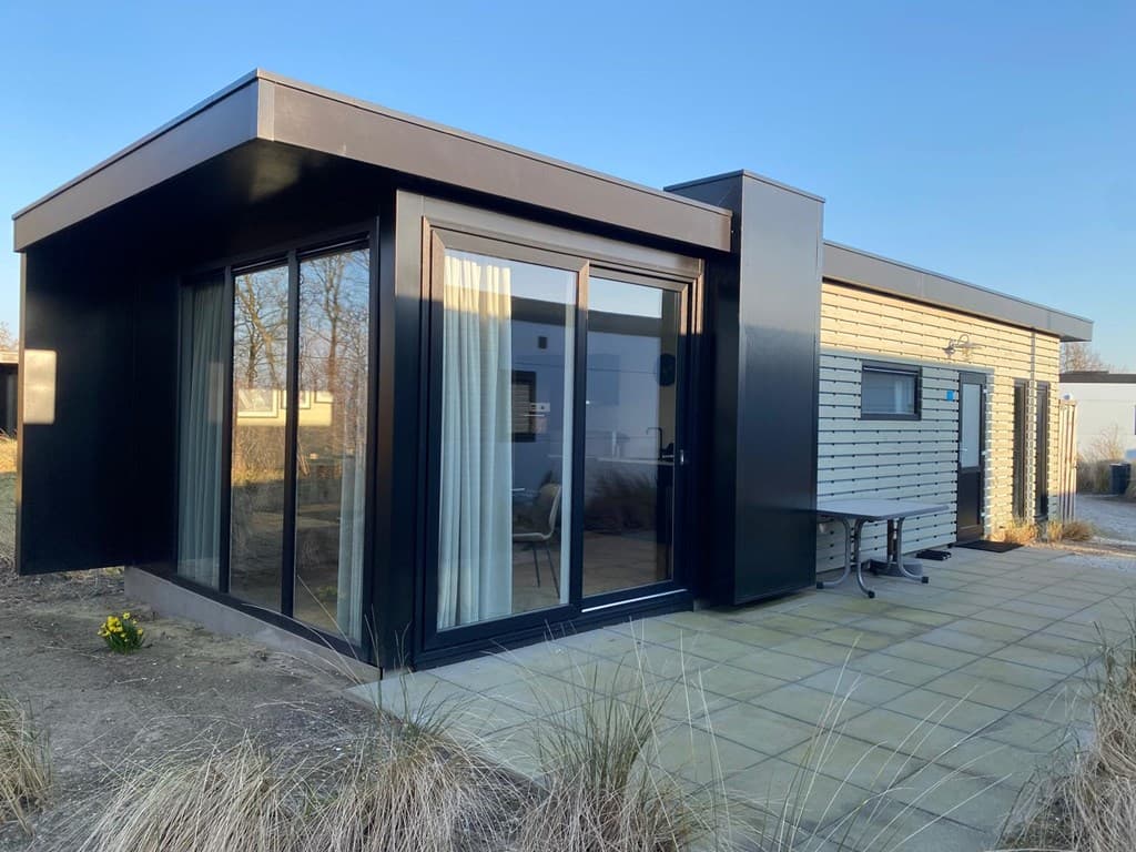 Moderne vakantiewoning met zwarte gevel, grote ramen en een betegeld terras, omringd door groen.