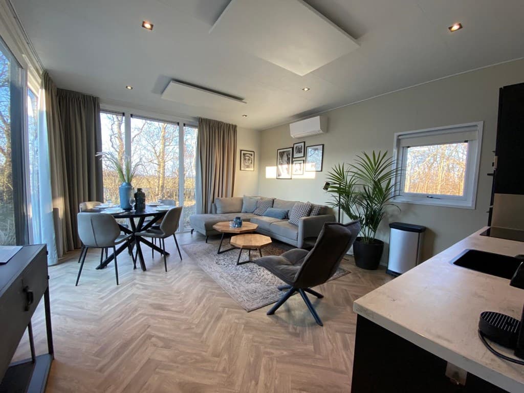 Woonkamer met banken, eettafel, en grote ramen met uitzicht op de natuur