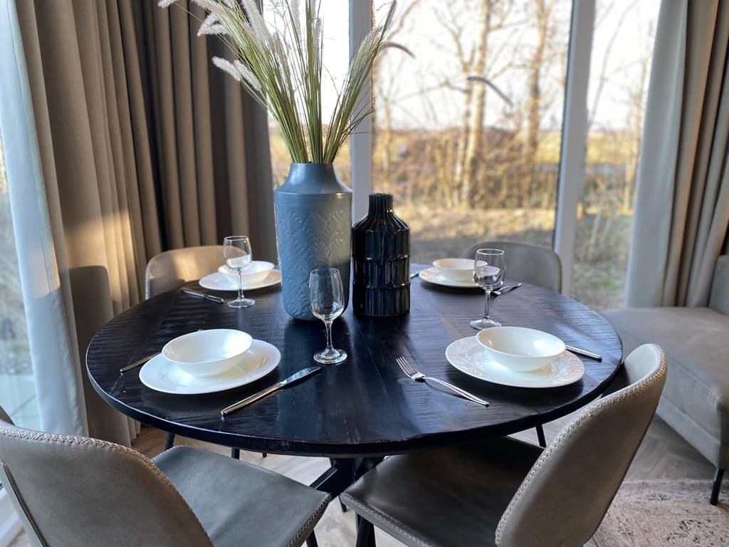 Eettafel met vier borden en glazen, vaas met bloemen op een ronde tafel bij grote ramen met uitzicht op groen.