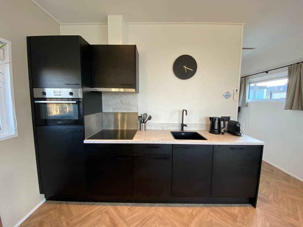Moderne keuken met zwarte kasten, oven, spoelbak en koffiezetapparaat, strakke afwerking met laminaatvloer.