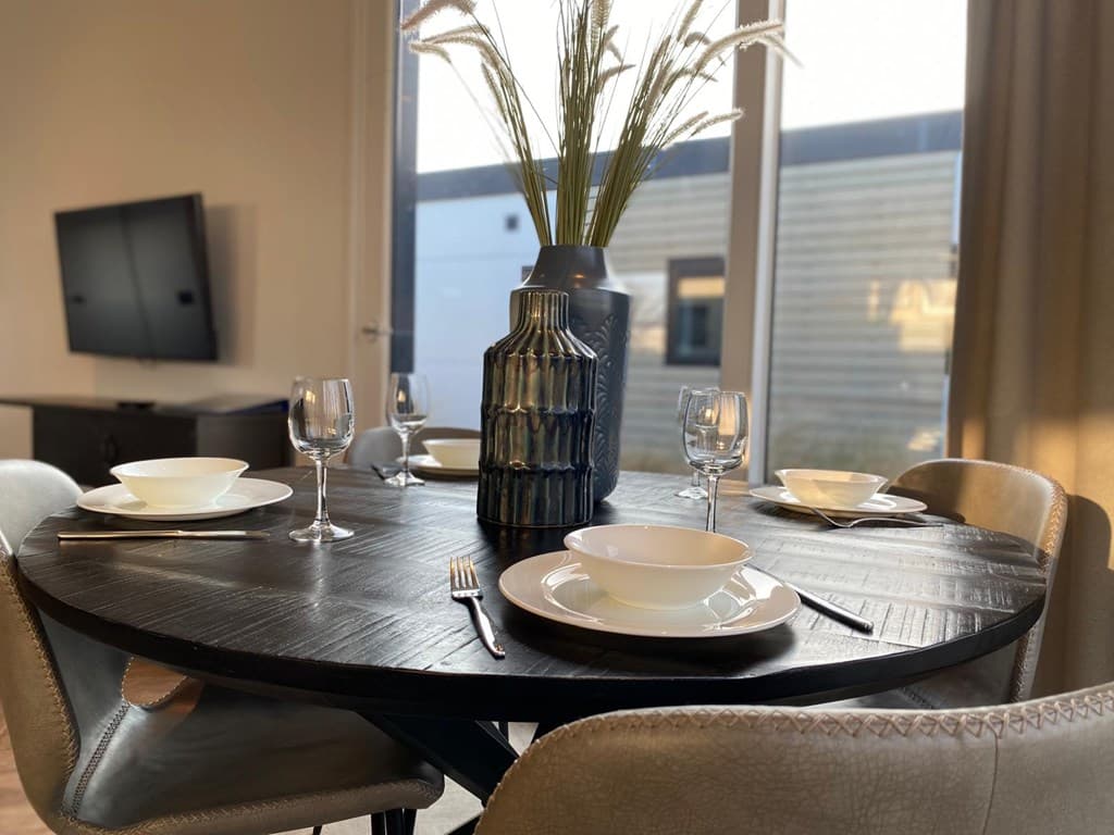 Eettafel met vier borden, glazen en een vaas met decoratieve takken in een lichte ruimte met grote ramen.
