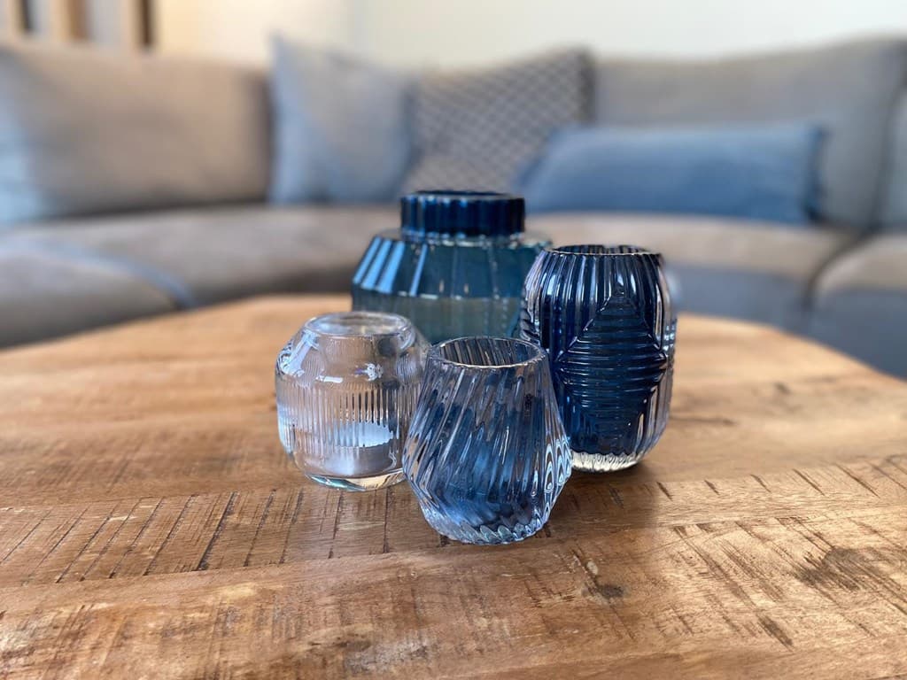 Een houten tafel met glazen vazen in verschillende blauw- en helder tonaliteiten, op de achtergrond een grijze bank.