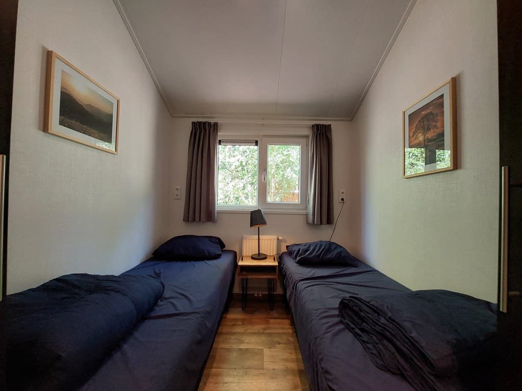 Twee eenpersoonsbedden met donkerblauwe dekbedden en een nachtkastje met lamp in een lichte kamer bij een raam.