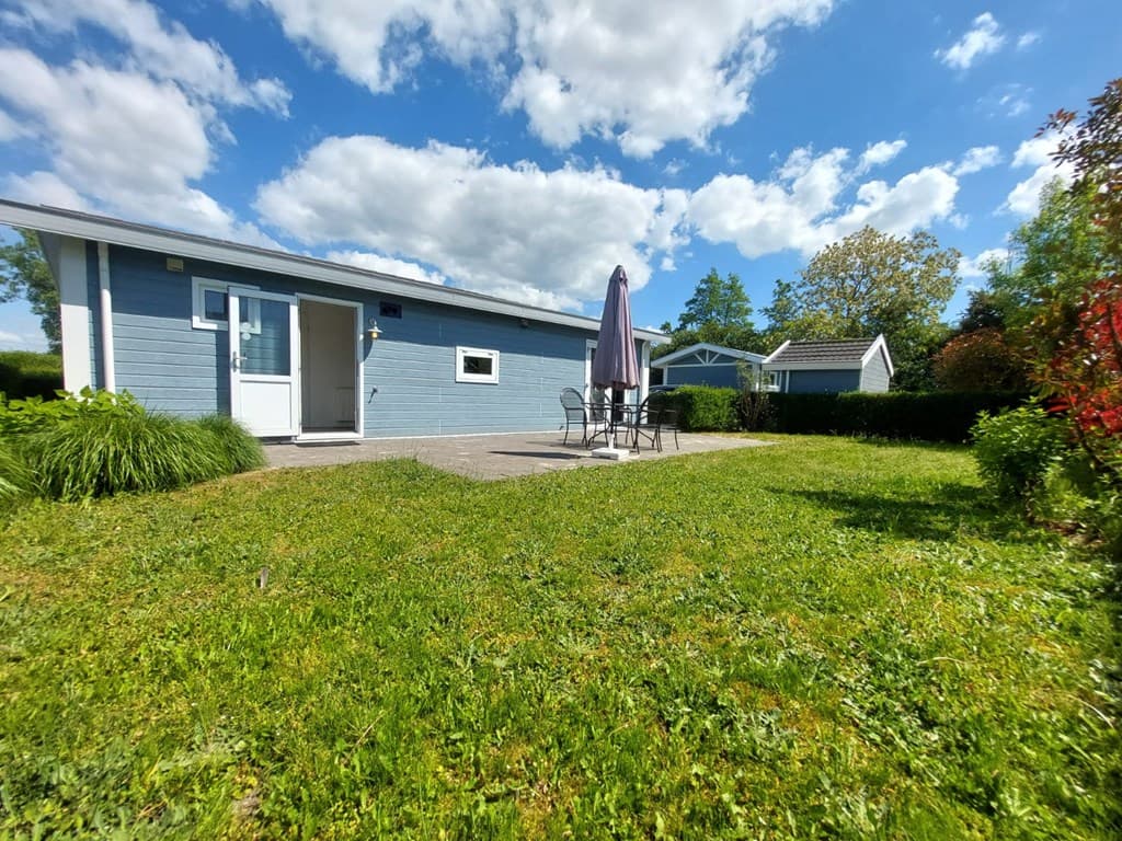 Buitenzicht op een vakantiewoning met terras, tuinmeubels en grasveld onder een blauwe lucht met wolken