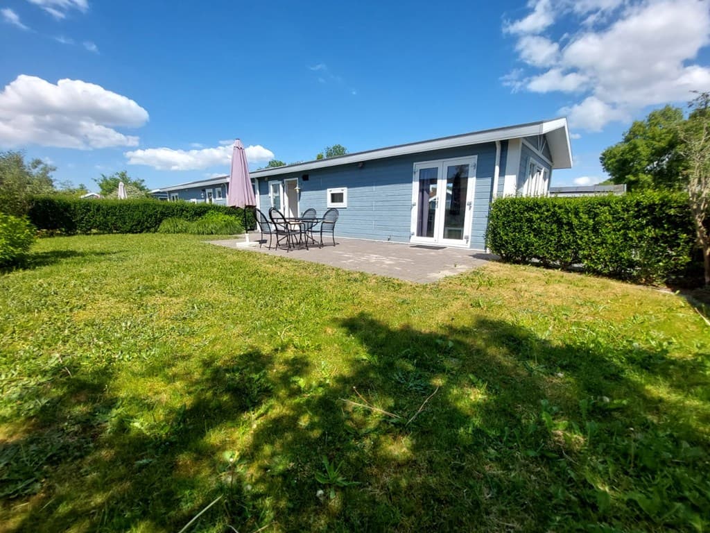 Tuin van een vakantiewoning met terras en tuinset, omringd door gras en heggen, onder een blauwe lucht met wolken