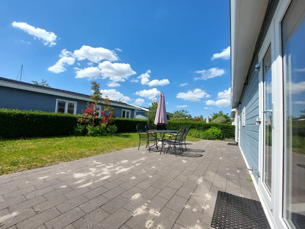 Terras met een tafel en stoelen, groen gazon en blauwe lucht met witte wolken achter een vakantiewoning
