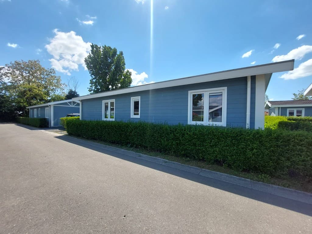 Zijaanzicht van een blauwe vakantiewoning met groene heggen en een heldere lucht met enkele wolken.