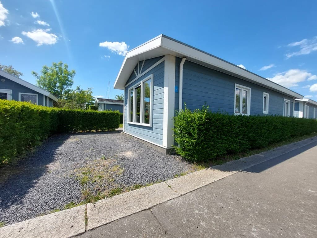 Een houten woning met blauwe gevel en grote ramen, omringd door groene hagen en grindpad.