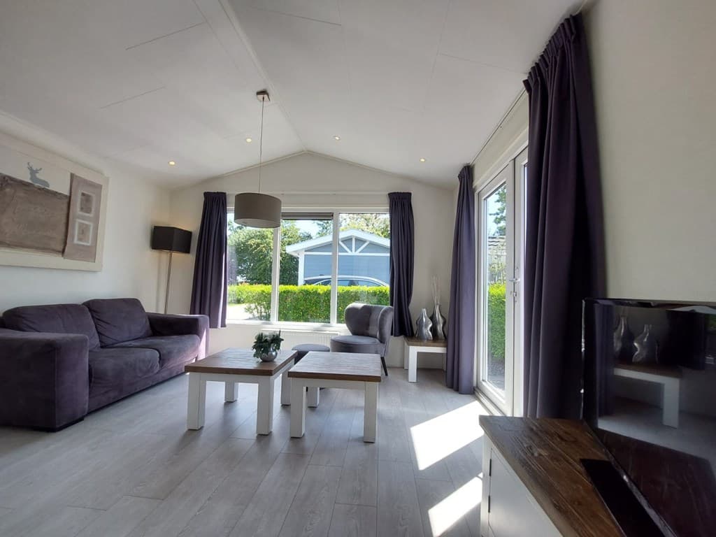 Woonkamer met zithoek, tafel en grote ramen die uitzicht bieden op de tuin en een blauwe woning buiten.