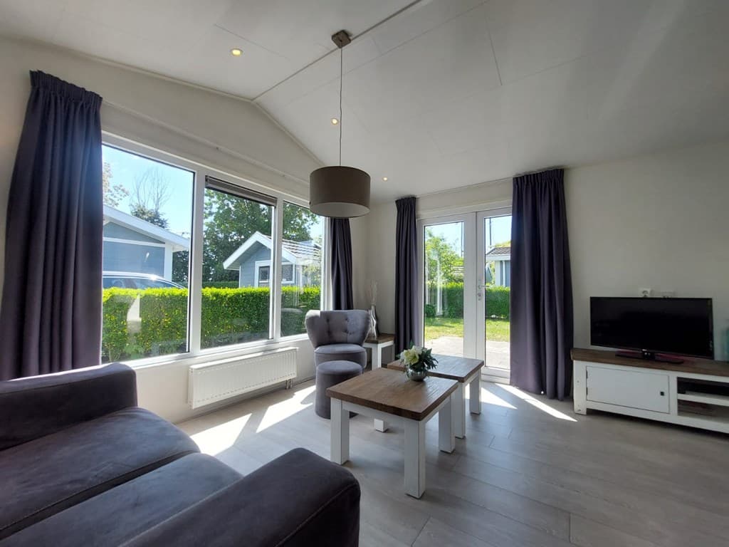 Lichte woonkamer met grote ramen, comfortabele fauteuils en een tv-meubel, omringd door groen buiten.