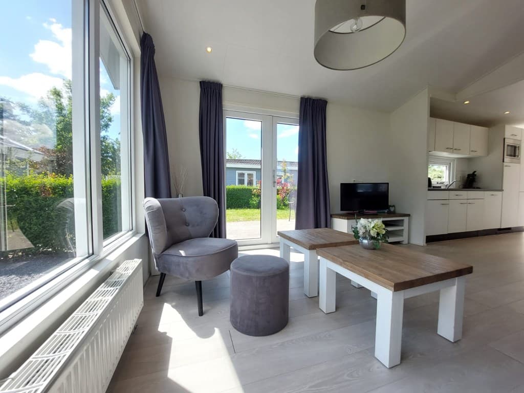Moderne woonkamer met grote ramen, een stoel en een poef, houten tafel en open keuken in EuroParcs Bad Hulckesteijn.