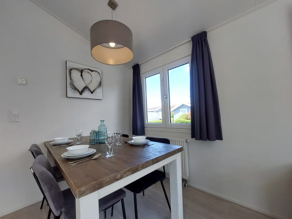 Eetkamertje met een houten tafel, vier stoelen en grote ramen met uitzicht op de buitenruimte en groene omgeving