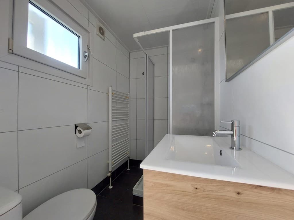 Badkamer met witte tegels, douchecabine, wastafel en toilet, radiator aan de muur en een raam voor natuurlijk licht