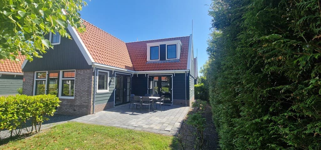Tuin met terrastegels en zitgelegenheid, groene heggen en een woning met een zadeldak en gevelbekleding in donkere kleur.