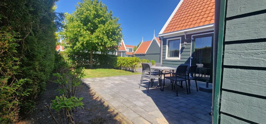 Tuin van een vakantiewoning met een terras, tuintafel en stoelen, omgeven door gras en heesters