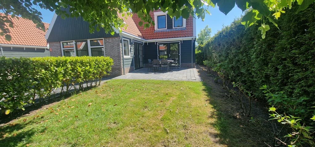 Buitenaanzicht van een woning met een betegeld terras en een groene tuin omgeven door hagen en struiken.