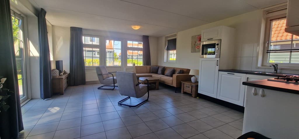 Lichte woonkamer met bank, stoelen, televisie en open keuken, grote ramen met uitzicht op buitenruimte