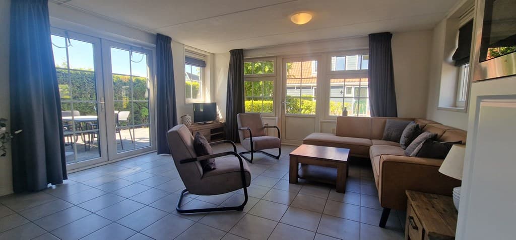 Zithoek met een bank, fauteuils en een tv, grote ramen bieden zicht op de tuin en het terras.