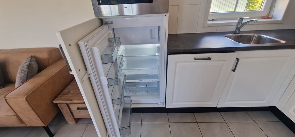 In de keuken staat een open koelkast met lege planken en een aanrecht met een gootsteen.