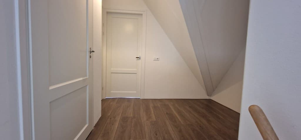 Lichte gang met een houten vloer, een deur naar een andere ruimte en schuine wanden.