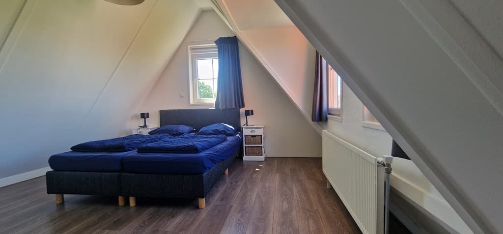 Slaapkamer met schuin dak, twee eenpersoonsbedden met donkerblauw beddengoed en een raam met gordijnen.