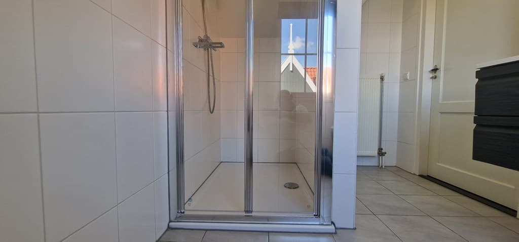Inloopdouche met glazen wand en tegelvloer, zichtbaar vanuit een hoek met de deur en radiator op de achtergrond