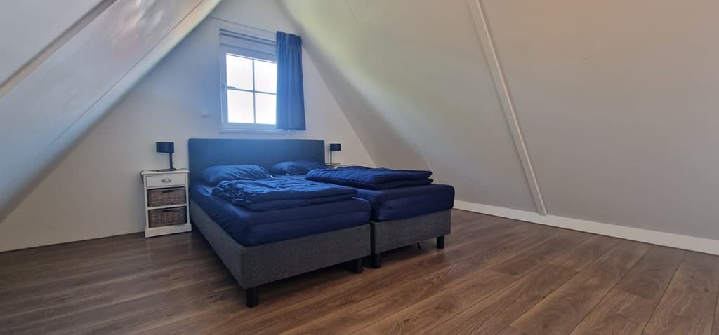 Slaapkamer met twee losse bedden, houten vloer en een raam met blauw gordijn onder een schuin plafond.