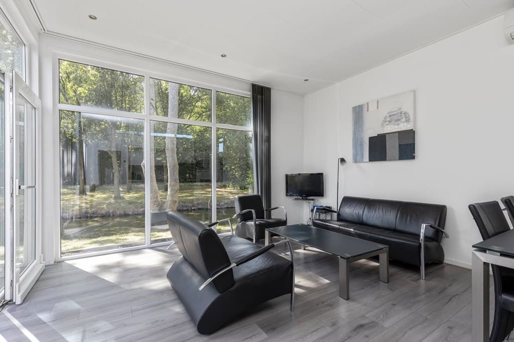 Licht en modern interieur met grote ramen, zithoek met zwarte banken en tv, uitzicht op groen buitengebied.