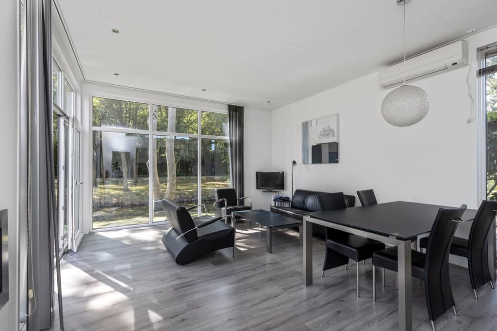 Moderne woonkamer met grote ramen, zwart leren meubilair en uitzicht op een groene tuin.