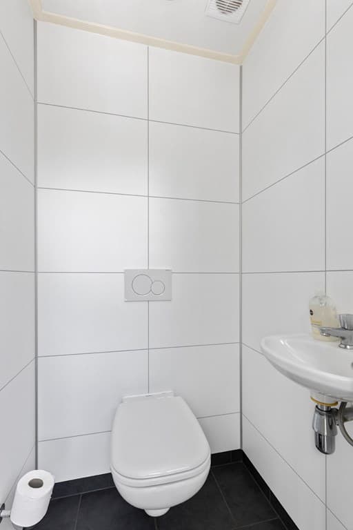 Witte toiletruimte met moderne tegels, een wastafel en een rol toiletpapier op de vloer.