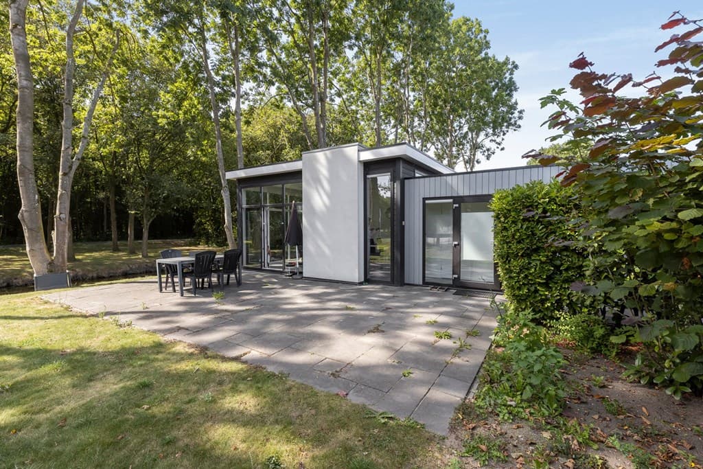 Moderne vakantiewoning met ruime terras en houten tuinmeubilair, omgeven door groene bomen en gras.