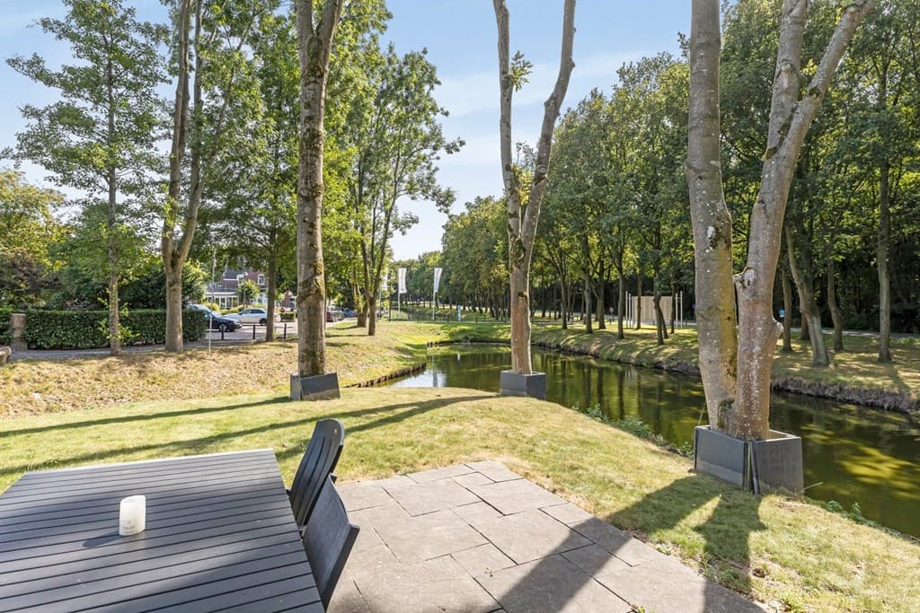 Uitzicht op een groene tuin met een waterloop en bomen, een terras met een tafel en stoelen in de foreground.