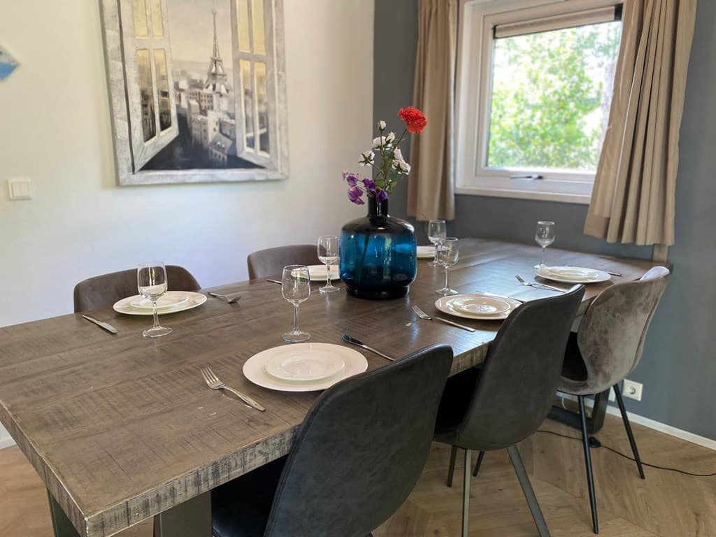 Eettafel met zes stoelen, vazen met bloemen en servies, grote ramen met uitzicht op groen buiten.