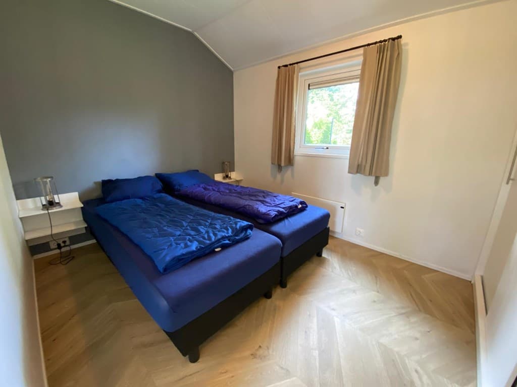 Bedkamer met twee losse bedden, blauwe dekbedden en raam met gordijnen. Houten vloer en neutrale wandkleur.