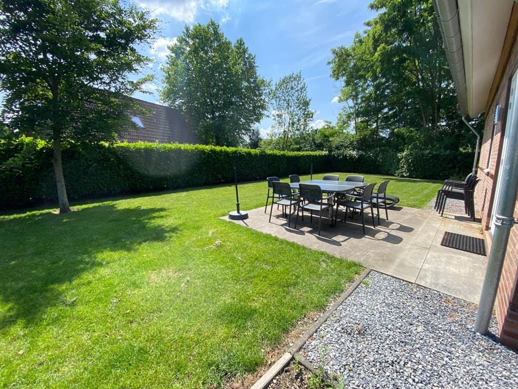 Tuin met gras en een betegeld terras met een grote tafel en stoelen, omringd door lage heggen en bomen.