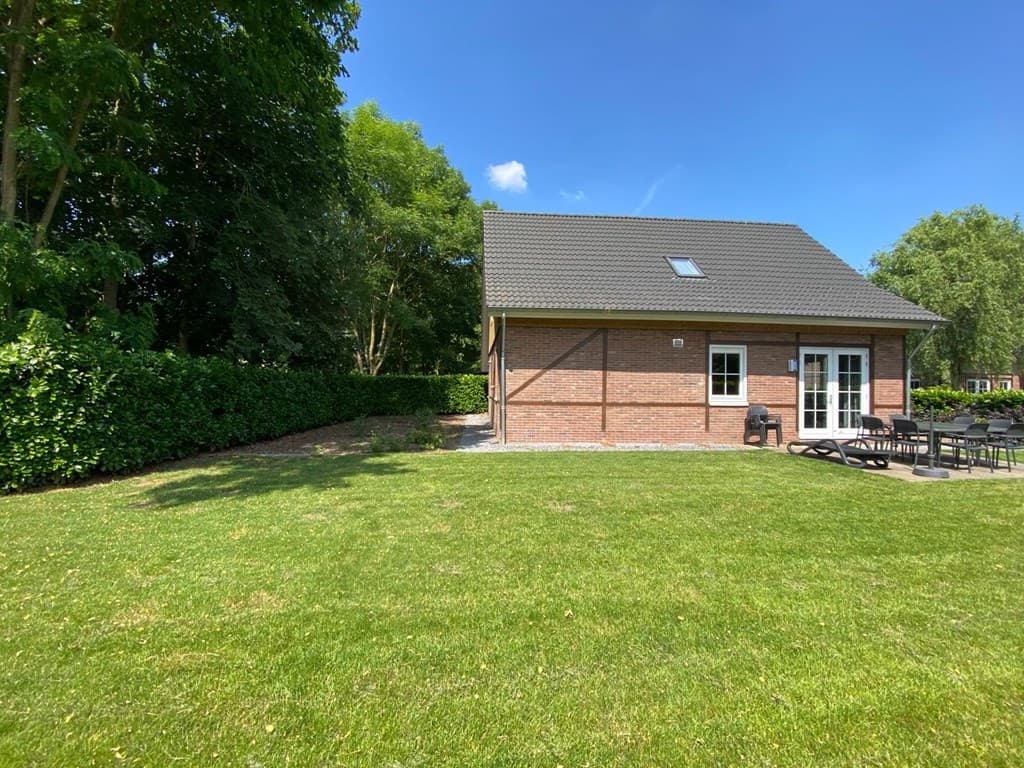 Vakantiewoning met een groene tuin en een stenen terras, omringd door hagen en bomen.