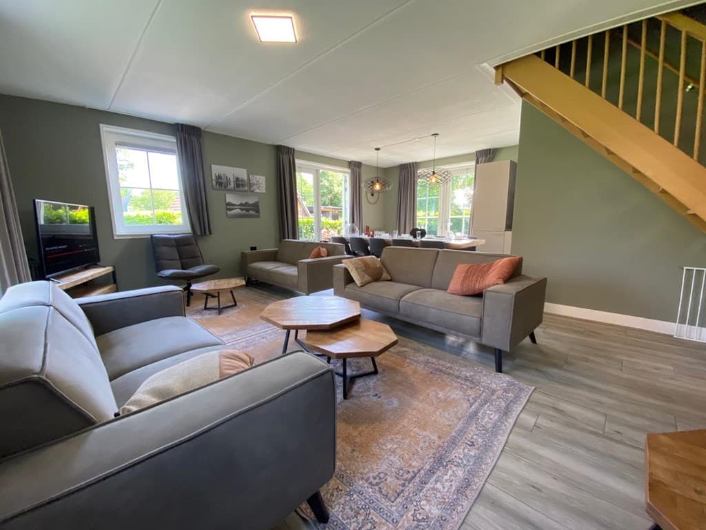 Ruime woonkamer met beige banken, houten tafels en een open trap, met grote ramen voor veel daglicht