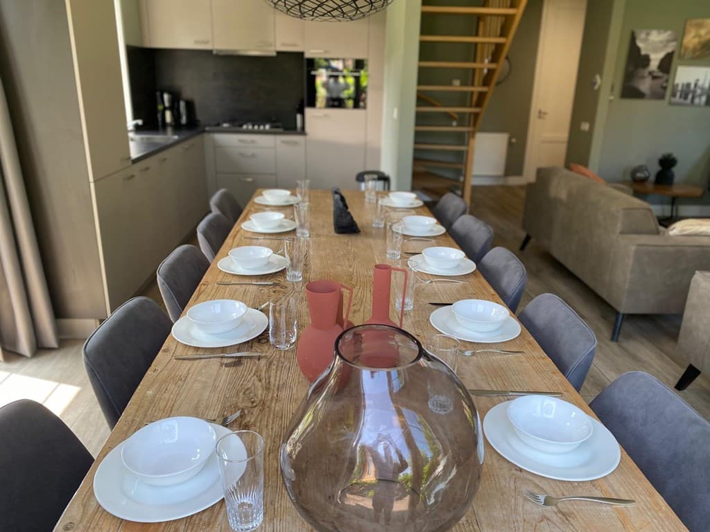 Eettafel met servies, glazen en decoratieve elementen in een open woonkamer met uitzicht op de keuken en trap.