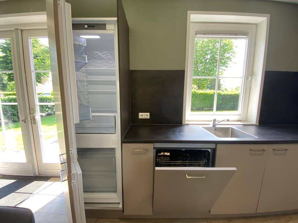 Moderne keuken met een open koelkast en een vaatwasser, grote ramen met uitzicht op groen buiten.