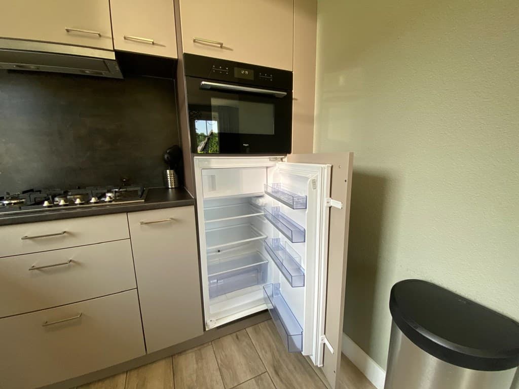 Open koelkast met transparante lades in moderne keuken, tegen een groene wand met een stalen afvalcontainer.