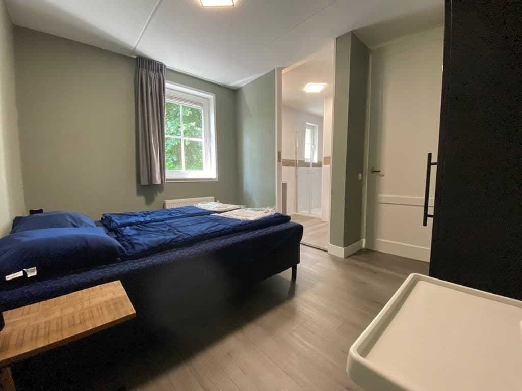 Slaapkamer met een bed, een nachtkastje en een raam met uitzicht op groene bomen.