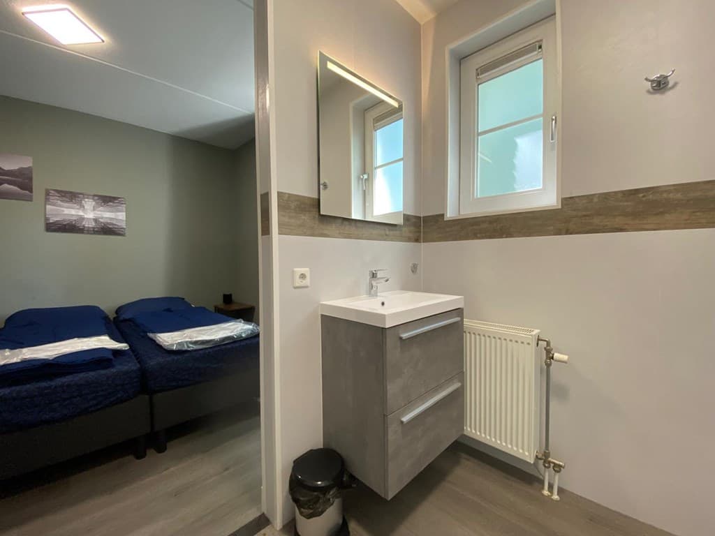 Moderne badkamer met wastafel en spiegellamp, naast twee eenpersoonsbedden en een klein raam.