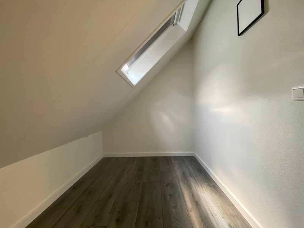 Ruimte met schuin plafond, houten vloer en een klein raam dat natuurlijk licht binnenlaat.