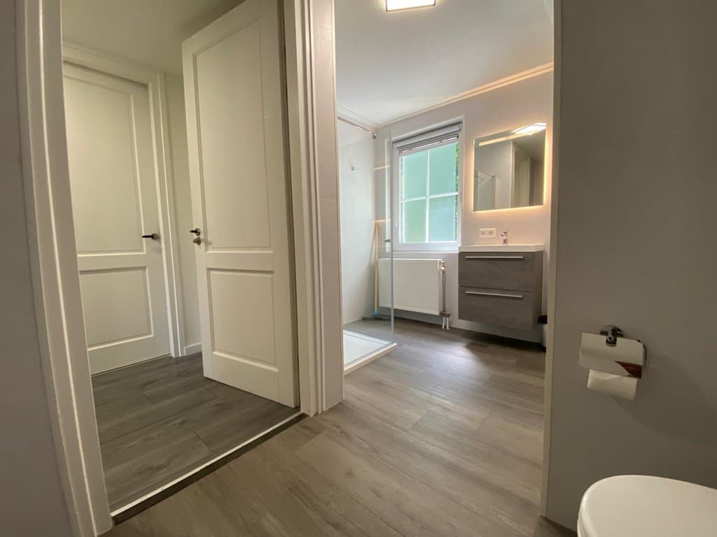 Moderne badkamer met grote raampartij, wastafel, douche en toegang tot een gang met deuren naar andere ruimtes