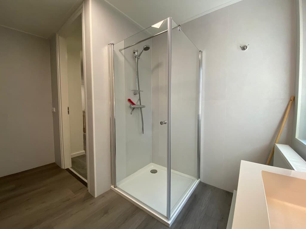 Douchehoek met glazen deuren, moderne douchekop en lichte wanden in een badkamer met houten vloer.