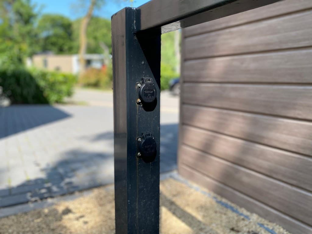 Zwarte metalen paal met twee elektrische aansluitingen en een asfaltpad op de achtergrond met bomen