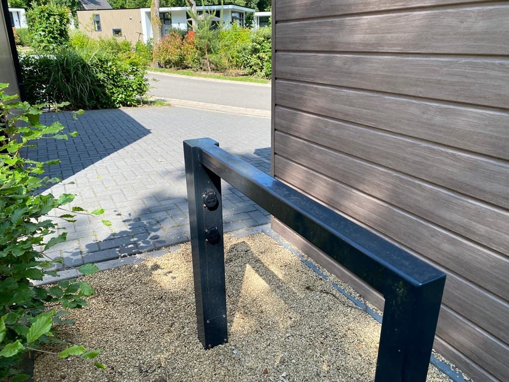 Zicht op een zwarte rek met een houten wand en grindgrond bij een vakantiehuis in een groene omgeving